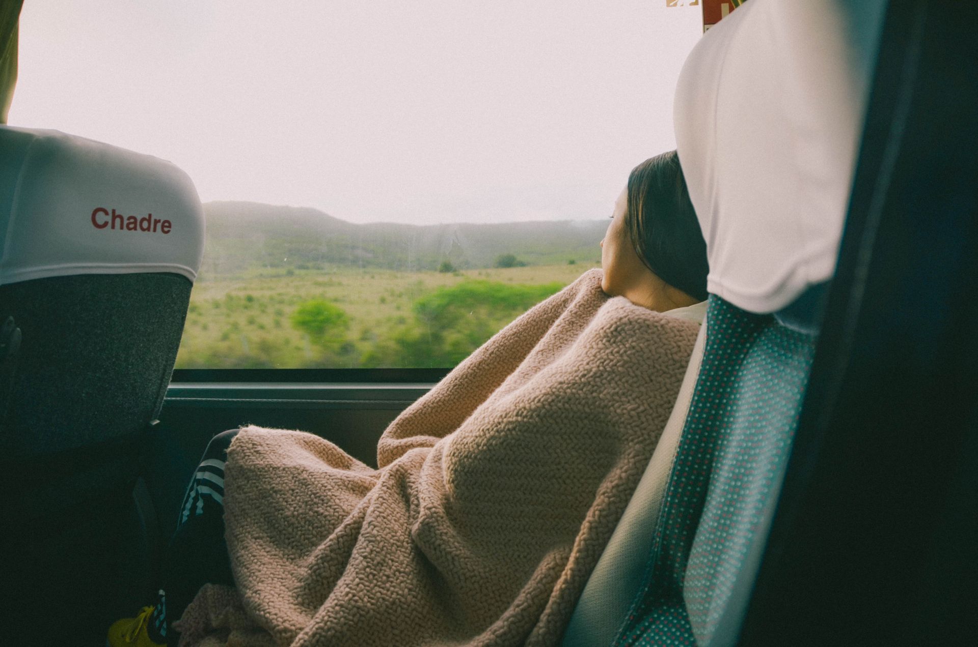 Pasajera disfrutando de un viaje cómodo en autobús, envuelta en una manta y contemplando el paisaje natural de Uruguay a través de la ventana.